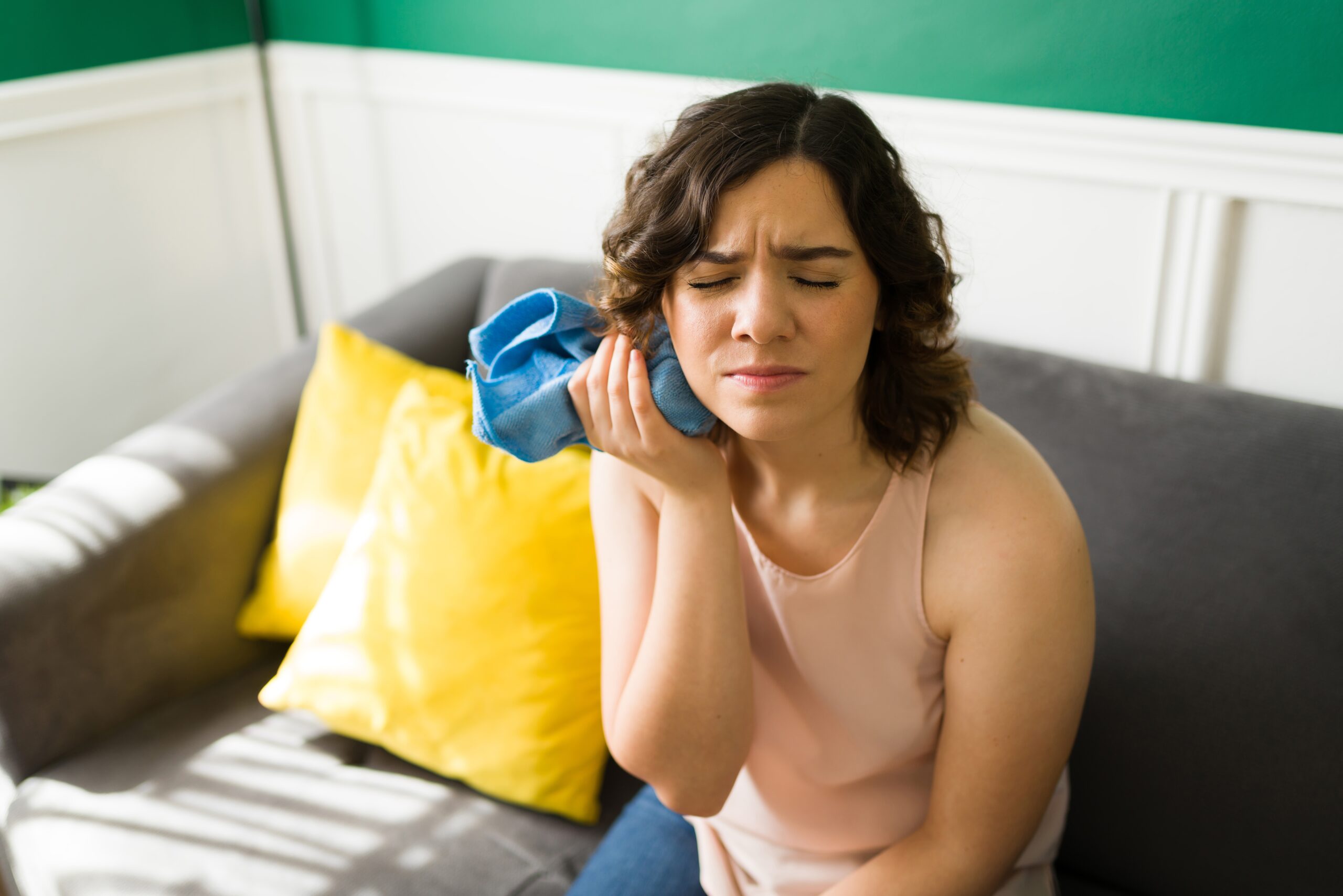 young woman sitting on a couch with an ice pack on her jaw, severe tooth pain